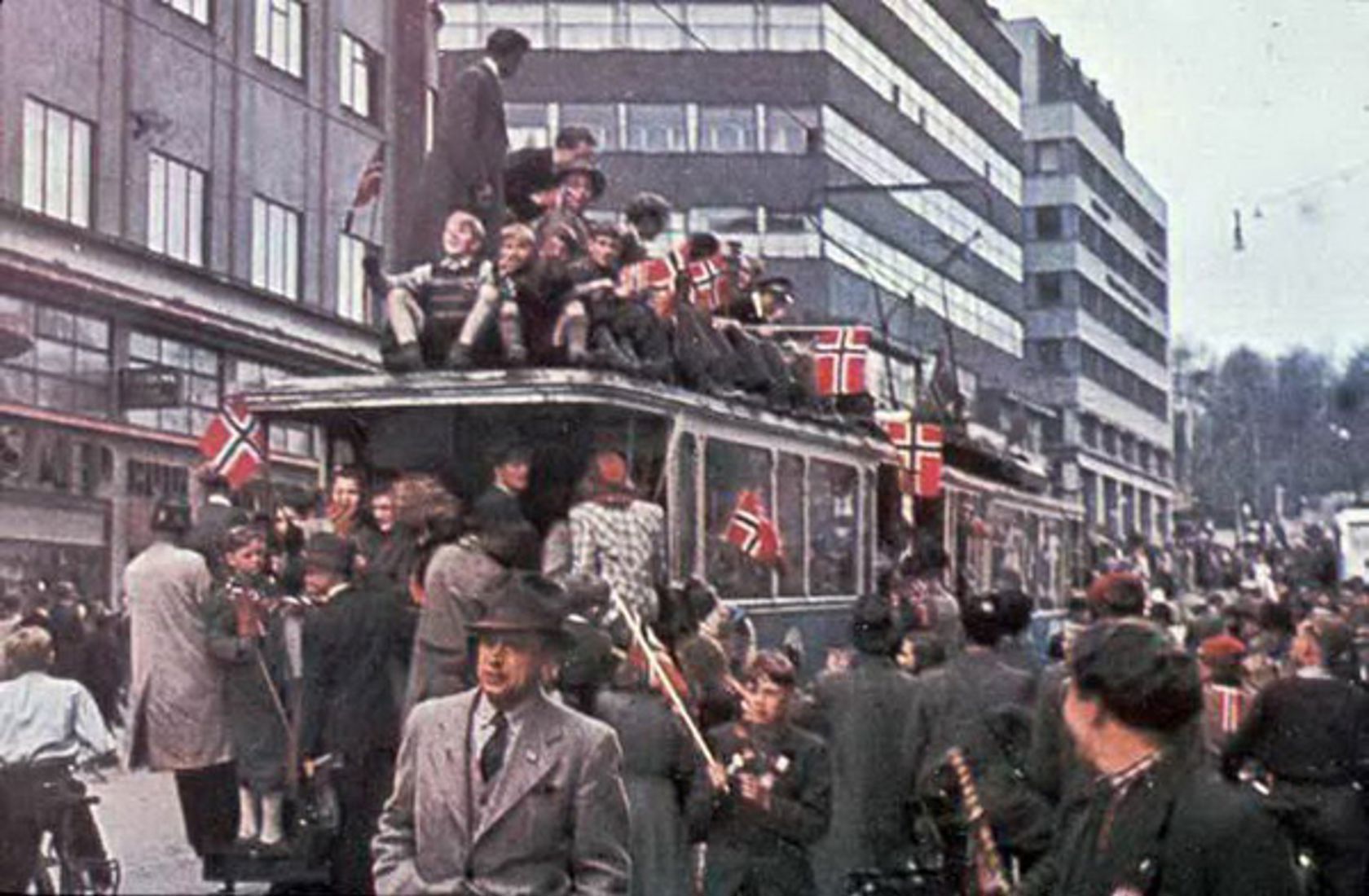 En folkemengde på og ved siden av trikken i dress og med norske flagg i hånden. 