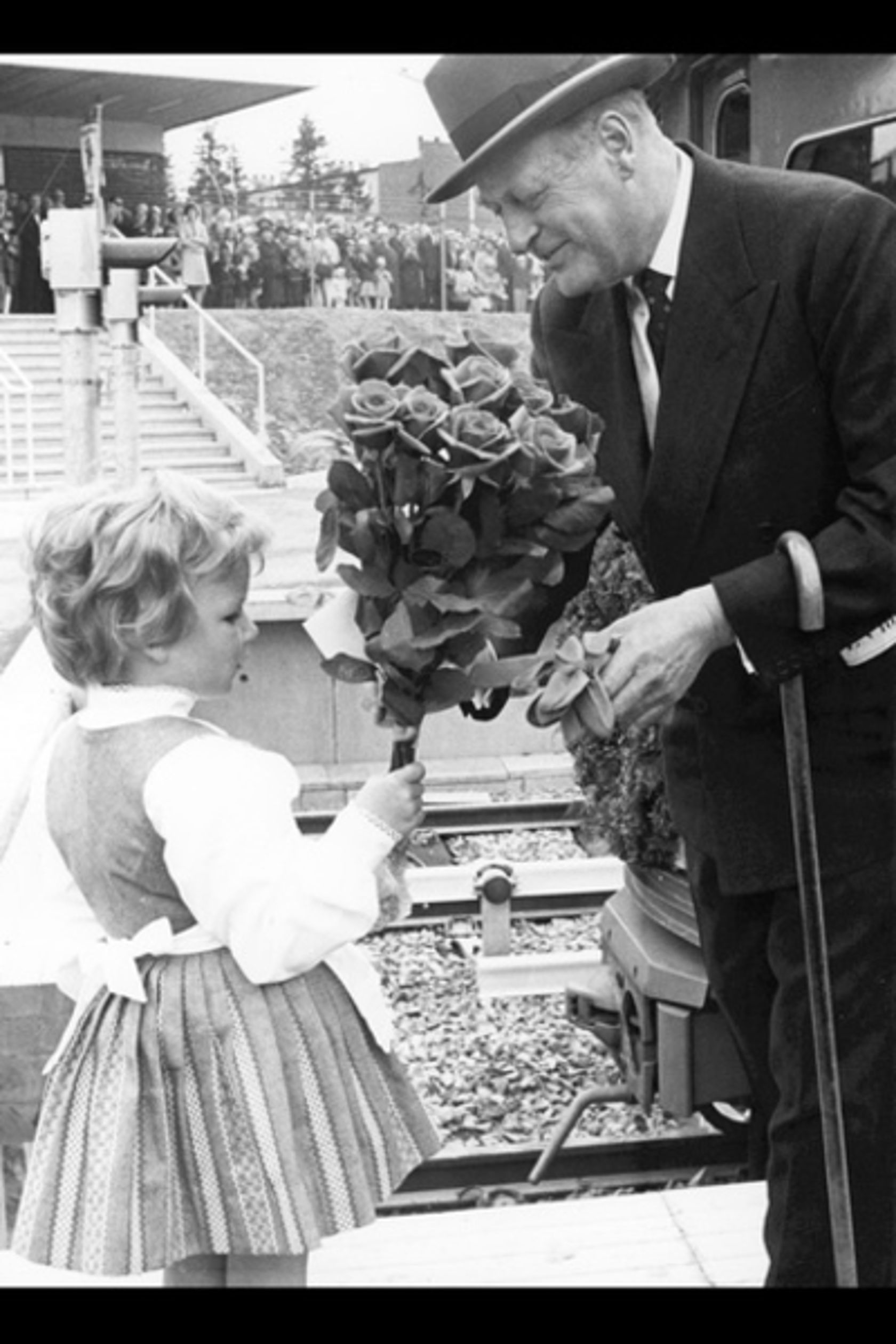 Svart-hvitt foto. Liten jente i pen kjole gir blomster til en eldre mann som smiler. 